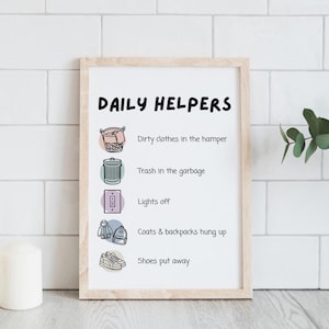May include: A framed print with the words "DAILY HELPERS" at the top. Below are illustrations and text: "Dirty clothes in the hamper," "Trash in the garbage," "Lights off," "Coats & backpacks hung up," and "Shoes put away." The frame is light brown.