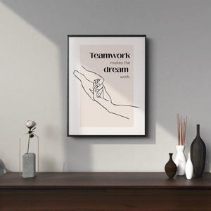 Op de afbeelding: Een ingelijste print met de tekst "Teamwork makes the dream work". Het kunstwerk toont een minimalistische lijntekening van twee handen die elkaar aanraken. De lijst is zwart en de achtergrond is lichtbeige. De print staat op een houten oppervlak.