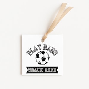 May include: Square white gift tag with the text "PLAY HARD" above a soccer ball graphic and "SNACK HARD" below. A tan ribbon is attached at the top. The text and soccer ball are in black and gray.