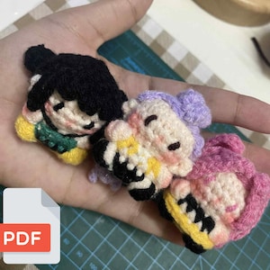 May include: Three handmade crochet doll pins are held in a hand. The dolls have black, purple, and pink hair, with yellow, green, and black accents. The dolls are smiling with closed eyes. The background is a green cutting mat and a beige and white checkered surface.