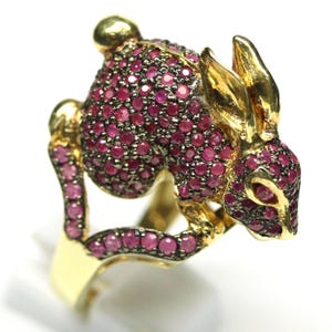 May include: A gold-toned ring shaped like a rabbit, featuring a body and head encrusted with numerous small, round, deep pink gemstones. The rabbit's ears, face, and tail are accented with polished gold metal, creating a detailed animal design.