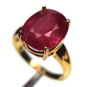 Könnte beinhalten: Ein Goldring mit einem großen, oval geschliffenen, tiefroten Edelstein, der von vier Krappen gehalten wird. Die Ringschiene ist glatt und poliert und reflektiert das Licht, während der facettierte Stein einen satten, tiefroten Farbton aufweist.