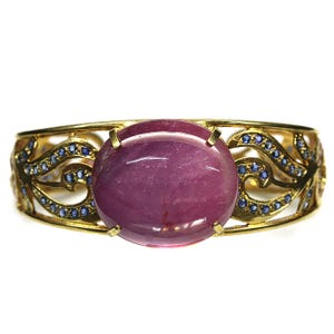 May include: A gold-toned bracelet featuring a large, oval, purplish-red gemstone at its center. The bracelet has an open design with intricate, swirling patterns and small blue gemstones. The setting is a bright, polished gold.