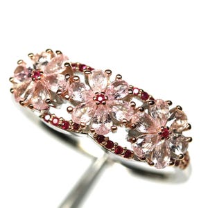 May include: A floral ring featuring pink gemstones arranged in a flower pattern. The ring has a silver band with rose gold accents and small red gemstones. The gemstones are set in a delicate, elegant design.