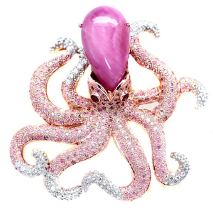 May include: A bejeweled octopus brooch with a large, oval, pink gemstone head. The tentacles are covered in pink gemstones, with silver-coloured gemstone tips. The brooch has a rose gold setting against a white background.