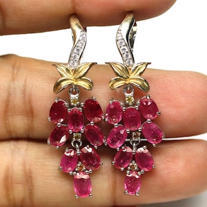May include: A pair of dangle earrings featuring clusters of oval-cut, deep pink gemstones. The earrings have gold-colored floral accents and silver-colored hooks with small, clear stones. The gemstones are arranged in a cascading design.