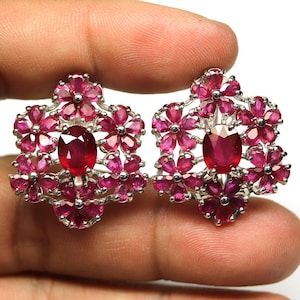 Peut inclure: Une paire de boucles d'oreilles argentées avec un motif floral. Chaque boucle d'oreille présente une grosse pierre précieuse rouge foncé de coupe ovale au centre, entourée de petites pierres précieuses roses en forme de pétales. Les boucles d'oreilles sont tenues par une main.