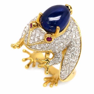 May include: A gold ring shaped like a frog, featuring a large oval blue sapphire cabochon on its back. The frog's body is encrusted with numerous small white diamonds, and it has two small red rubies set as eyes, with gold legs and feet.