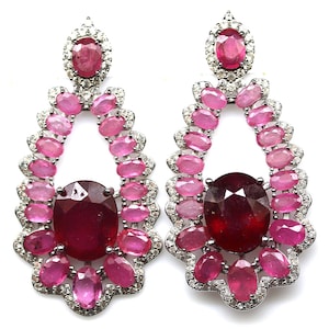 May include: A pair of silver earrings featuring a large oval ruby at the center, surrounded by smaller pink and ruby-colored stones. The earrings have a teardrop shape and are set with small, clear accent stones.