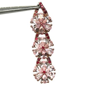 Peut inclure: Boucle d'oreille délicate en forme de fleur avec une finition or rose. La boucle d'oreille présente trois grappes en forme de fleur, chacune composée de pierres précieuses roses claires et rouges. La boucle d'oreille est tenue par un outil en métal argenté sur fond blanc.