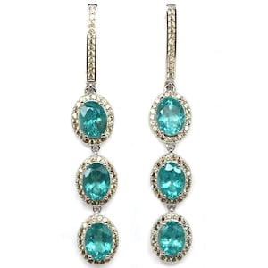 May include: A pair of dangling earrings with three oval, teal-colored gemstones. Each gemstone is surrounded by a halo of smaller, light-colored stones. The earrings have a silver-colored metal setting and a post-and-clip closure.