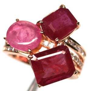 May include: A rose gold ring featuring three gemstones: a round pink stone and two rectangular, deep red rubies. The ring is accented with small, clear stones along the band, creating a luxurious and eye-catching design.