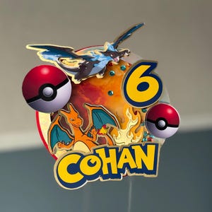 May include: A vibrant cake topper with a Charizard and a blue and black dragon design. The topper includes two red and white Pokeball designs, the number 6, and the name "COHAN" in blue and gold lettering. The background has an orange and red design.