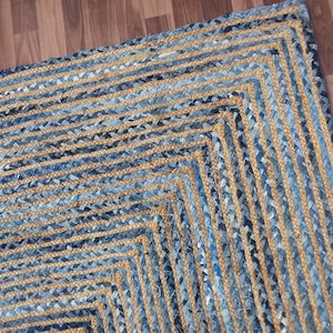 May include: Braided jute rug with a geometric design. The rug features alternating rows of natural tan and blue denim-like material, creating a textured, eye-catching pattern. The rug's edges are defined by a series of angled lines, adding visual interest to the floor covering.