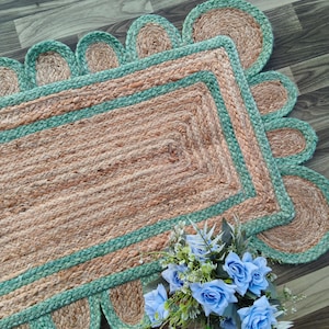May include: A rectangular jute rug with a scalloped edge and a teal border. The rug has a natural, woven texture and is decorated with a bouquet of blue roses. The rug's dimensions are not visible in the image.