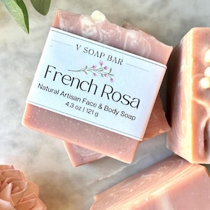 May include: A rectangular bar of pink soap with white swirls, wrapped in a white paper label that reads V SOAP BAR French Rosa Natural Artisan Face & Body Soap 4.3 oz | 121 g. The soap has a smooth texture and a light pink hue throughout the bar.