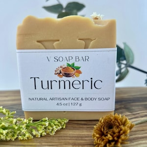 May include: A rectangular light tan soap bar with a white paper label. The label features the text V SOAP BAR, Turmeric, and NATURAL ARTISAN FACE & BODY SOAP, along with an illustration of turmeric root and powder. The label states 4.5 oz | 127 g.