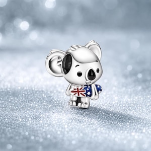 Op de afbeelding: Een zilverkleurige metalen bedel in de vorm van een koala. De koala heeft grote oren, zwarte ogen en een zwarte neus. Het dier houdt een klein surfboard vast dat is versierd met de blauwe, witte en rode kleuren van de Australische vlag.
