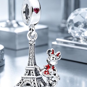 Puede incluir: Un charm colgante de plata de ley con una torre Eiffel detallada y una peque&ntilde;a figura con un vestido rojo de lunares blancos. El charm incluye una anilla con el sello S925 y una peque&ntilde;a piedra roja en forma de coraz&oacute;n en la parte superior.