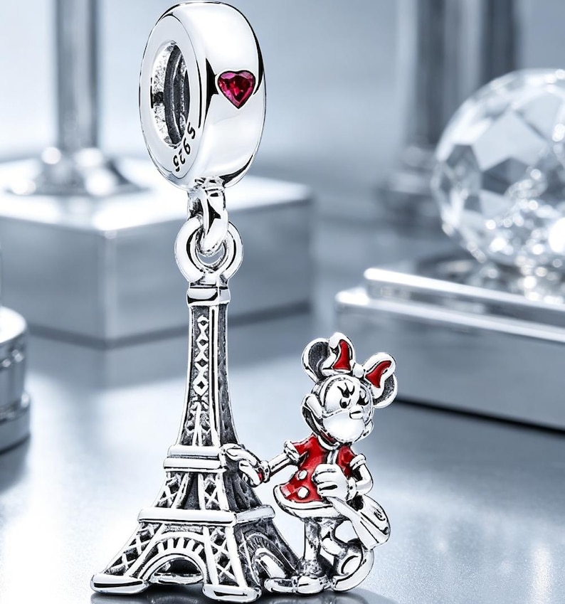 Puede incluir: Un charm colgante de plata de ley con una torre Eiffel detallada y una peque&ntilde;a figura con un vestido rojo de lunares blancos. El charm incluye una anilla con el sello S925 y una peque&ntilde;a piedra roja en forma de coraz&oacute;n en la parte superior.