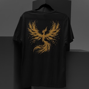 May include: Black t-shirt with a gold phoenix design on the back. The phoenix is shown with outstretched wings and a flowing tail, with a splatter effect. The shirt is hanging on a black hanger.