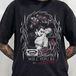 Model with a dark bob haircut and tattooed arms, standing against a metallic industrial wall, wearing a black snow-washed oversized t-shirt. The t-shirt features a large gothic-style graphic print in white, black, and deep red. The print includes an illustration of a vampire, a pair of skeletal hands holding a heart, a close-up of a vampire teeth, all framed by a border of thorny vines and dark roses. Below the graphic, the text reads "WILL YOU BE MY VALENTINE?" in a mix of white and red font
