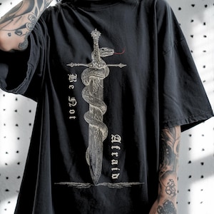 May include: Dark gray t-shirt featuring a detailed graphic of a sword entwined by a snake. The design includes the words "Be Not Afraid" in a gothic font. The shirt has a classic crew neck and short sleeves.