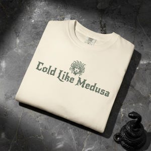 Folded ivory Comfort Colors t-shirt with dark green gothic-style lettering that says “Cold Like Medusa,” featuring a Medusa head graphic above the text.
