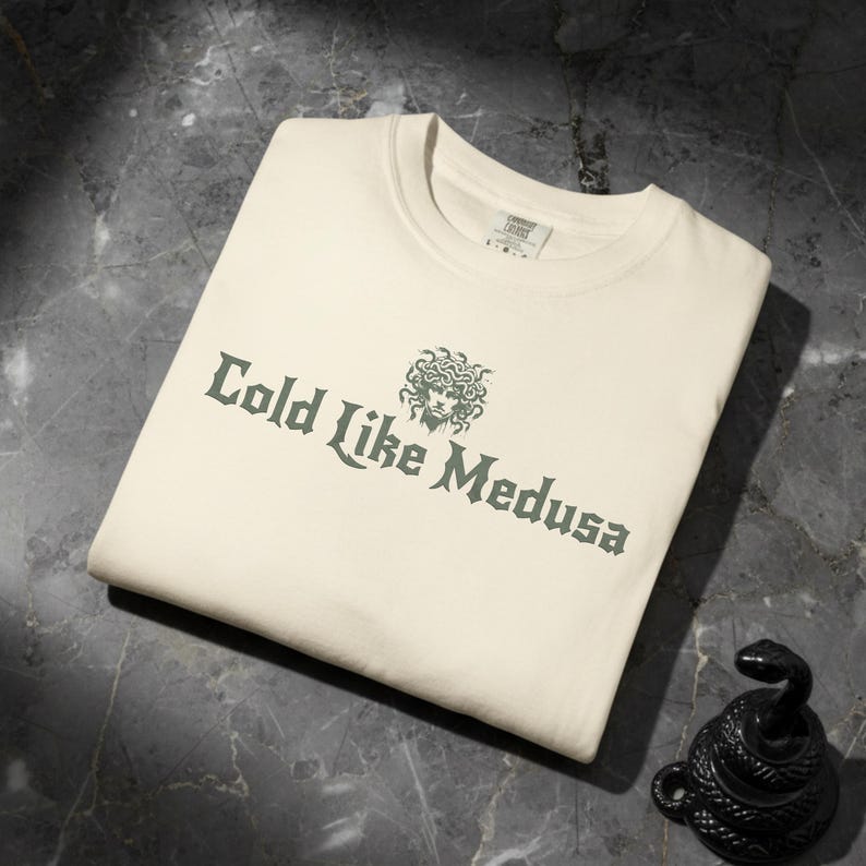 Folded ivory Comfort Colors t-shirt with dark green gothic-style lettering that says “Cold Like Medusa,” featuring a Medusa head graphic above the text.