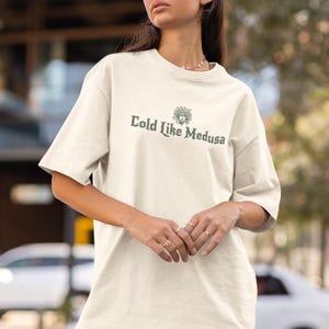 May include: Cream-colored t-shirt with the text "Cold Like Medusa" in a dark green font. The shirt features a graphic of Medusa's head above the text. The t-shirt has short sleeves and a crew neck.