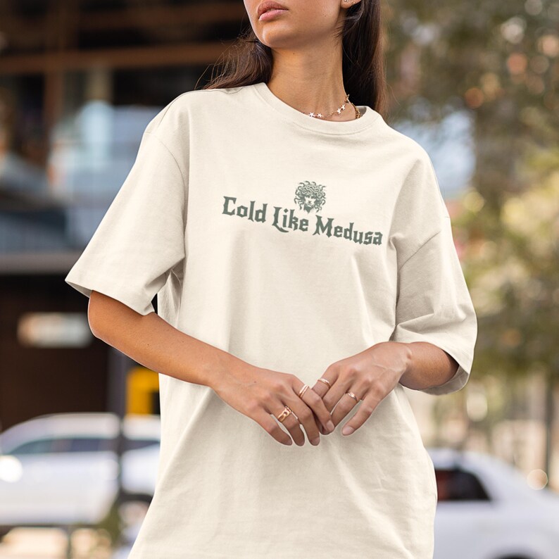 May include: Cream-colored t-shirt with the text "Cold Like Medusa" in a dark green font. The shirt features a graphic of Medusa's head above the text. The t-shirt has short sleeves and a crew neck.