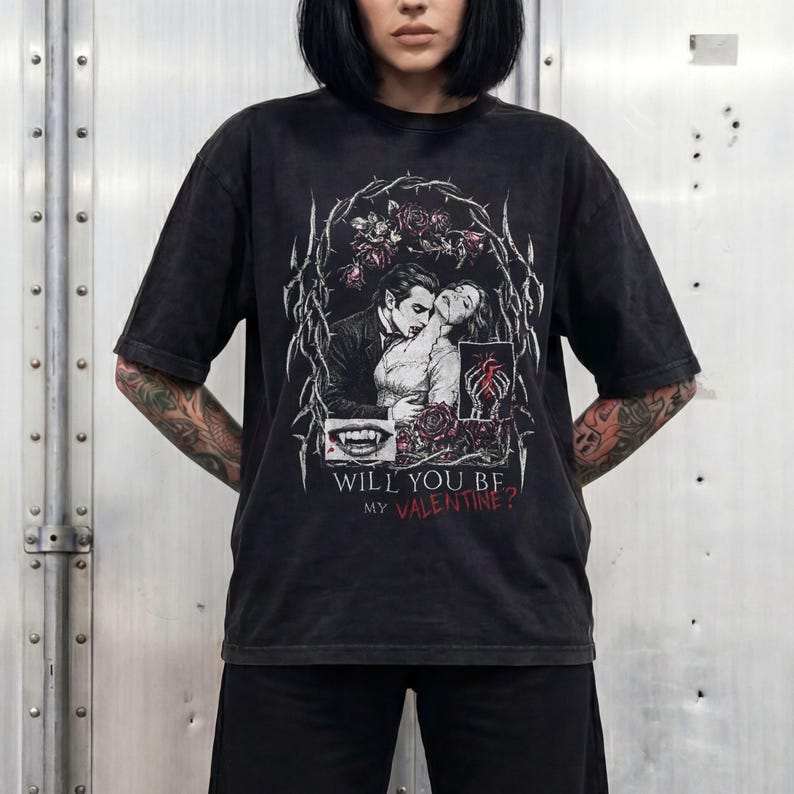 Model with a dark bob haircut and tattooed arms, standing against a metallic industrial wall, wearing a black snow-washed oversized t-shirt. The t-shirt features a large gothic-style graphic print in white, black, and deep red. The print includes an illustration of a vampire, a pair of skeletal hands holding a heart, a close-up of a vampire teeth, all framed by a border of thorny vines and dark roses. Below the graphic, the text reads "WILL YOU BE MY VALENTINE?" in a mix of white and red font
