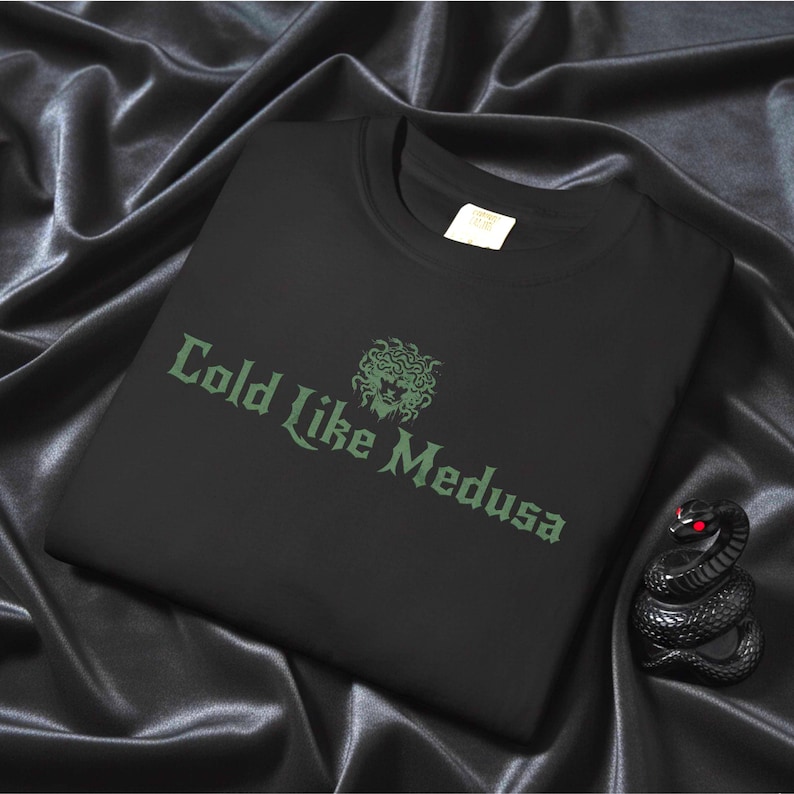 Folded black Comfort Colors t-shirt on black satin fabric with gothic-style lettering that reads “Cold Like Medusa” in dark green, above a Medusa head graphic. A black snake figurine with red eyes is placed beside the shirt.
