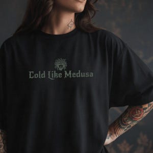 Person wearing a black Comfort Colors t-shirt with dark green gothic-style lettering that says “Cold Like Medusa,” featuring a Medusa head graphic above the text.