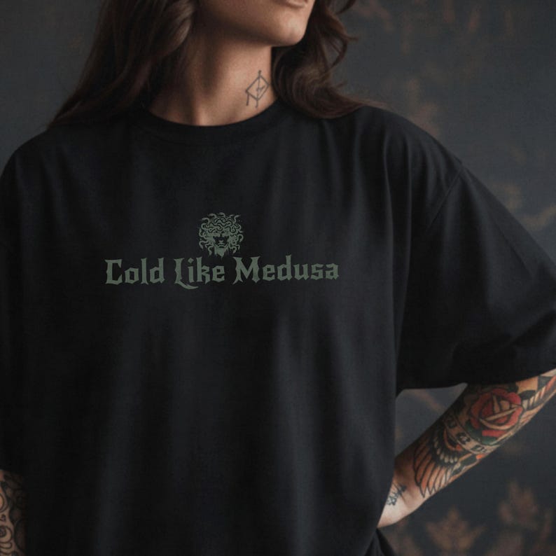 Person wearing a black Comfort Colors t-shirt with dark green gothic-style lettering that says “Cold Like Medusa,” featuring a Medusa head graphic above the text.