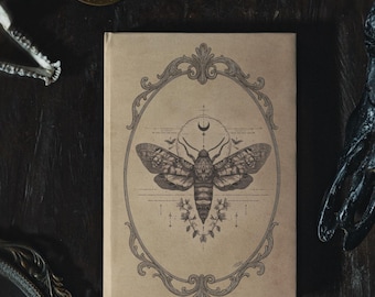 May include: A tan-colored journal featuring a detailed illustration of a moth within an ornate oval frame. The moth design includes a crescent moon and floral accents. The journal is set on a dark, textured surface.