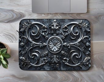 Puede incluir: Funda rectangular para portátil con un diseño plateado en relieve sobre un fondo gris oscuro. El diseño presenta patrones arremolinados, elementos florales y un motivo circular central. Un portátil es parcialmente visible en la parte superior.