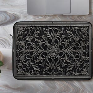 Op de afbeelding: Zwarte laptoptas met een sierlijk, zilverkleurig bloemmotief. De rechthoekige hoes heeft een ritssluiting en een symmetrisch patroon. Een laptop is gedeeltelijk zichtbaar op de achtergrond.