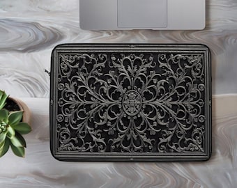 Puede incluir: Una funda rectangular para portátil con fondo negro y un patrón de filigrana plateado en relieve detallado. El diseño consta de volutas simétricas intrincadas y motivos florales centrados alrededor de un emblema ovalado.