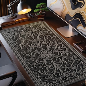 Puede incluir: Una alfombrilla de escritorio gris oscuro con un diseño floral en relieve ornamentado. La alfombrilla es rectangular y presenta un patrón simétrico detallado. Una lámpara de escritorio, un bonsái y un monitor de ordenador son visibles.