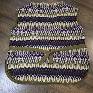 May include: A sleeveless knit vest with a geometric pattern in cream, purple, olive green, and navy blue. The vest has a matching olive green tie belt and trim. The overall design is a classic, vintage style.