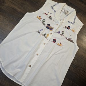 May include: White sleeveless button-down shirt with embroidered desert landscape design. The embroidery features cacti, mountains, and pottery in various colors. The collar and cuffs have a decorative blue stitching.