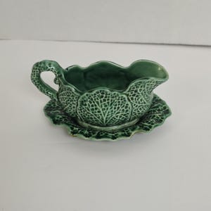 May include: A green ceramic gravy boat with an attached saucer, featuring a textured cabbage leaf pattern throughout the exterior. The piece has a curved handle and a scalloped rim, finished with a glossy glaze that highlights the intricate veins.