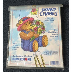 May include: A Dimensions counted cross stitch kit for wind chimes featuring a brown bear wearing a pink hat and blue overalls while holding a bouquet of flowers. The package includes text stating 10 inches long with chimes and kit contents.