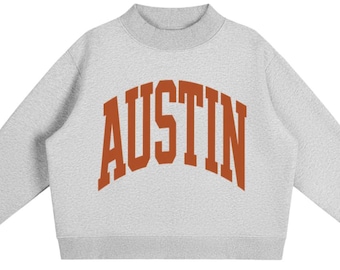 May include: A light grey long-sleeve sweatshirt with a mock neck collar. The front features the word AUSTIN printed in a large, arched, burnt orange collegiate-style font. The garment has ribbed cuffs and a ribbed hem, providing a structured fit.