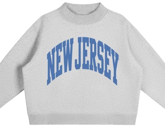 May include: A light grey crewneck sweatshirt featuring a mock neck collar and long sleeves. The text NEW JERSEY is printed across the chest in a bold, arched, blue collegiate-style font. The garment has ribbed cuffs and a ribbed hemline.