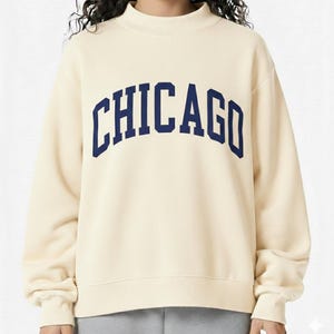 May include: Cream-colored sweatshirt with a mock neck and long sleeves. The word "CHICAGO" is printed across the chest in navy blue, arched lettering. The sweatshirt is a casual style.
