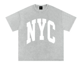 Op de afbeelding: Lichtgrijs t-shirt met de witte letters "NYC" in een boogvormig ontwerp op de voorkant. Het shirt heeft een ronde hals en korte mouwen. De stof heeft een licht vervaagde, vintage look.