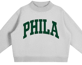May include: A light grey long-sleeve sweatshirt with a mock neck collar and ribbed cuffs. The front features the word PHILA printed in a large, dark green, arched collegiate-style font across the chest area, centred on the heathered fabric.