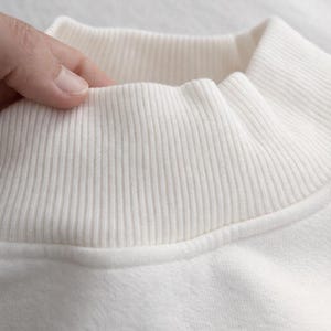 May include: Close-up of a cream-colored sweatshirt collar. The ribbed collar is a lighter shade of cream than the body of the sweatshirt. The fabric appears soft and comfortable. The collar is being touched by a hand.